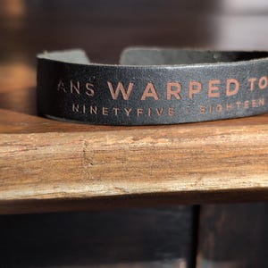 May include: Black leather cuff bracelet with embossed text reading "ANS WARPED TOUR NINETYFIVE EIGHTEEN". The bracelet is curved and sits on a wooden surface. The text is in a contrasting brown color.