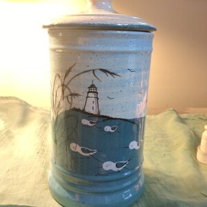 May include: A white ceramic jar with a blue and white lighthouse scene. The jar has a lid and is decorated with a lighthouse, seagulls, and grass.