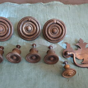 May include: A collection of antique brass cabinet hardware, including round knobs with intricate designs, a pull handle with a decorative floral design, and smaller round knobs with a raised center.