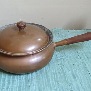 May include: A copper saucepan with a wooden handle and a copper lid. The saucepan has a rounded bottom and a flared rim. The lid has a wooden knob.