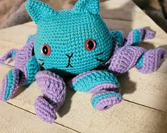 May include: A handmade turquoise and purple crochet cat-topus plush toy. The cat's head has pointed ears, pink button eyes, and a stitched face. The tentacles are purple with turquoise stripes. The toy is on a white wooden surface.