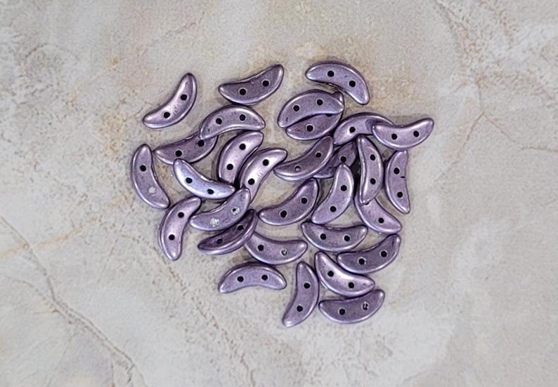 May include: A pile of small, crescent-shaped beads in a light purple color with a metallic sheen.