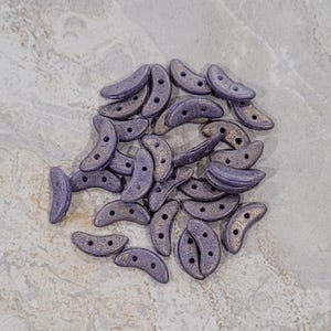 May include: A pile of small, crescent-shaped beads in a deep purple color with a subtle shimmer. The beads have two holes in each.
