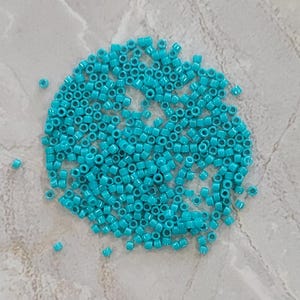May include: A collection of small, turquoise seed beads scattered on a light gray surface. The beads are cylindrical with a hole running through the center, ideal for stringing into jewelry or other crafts.