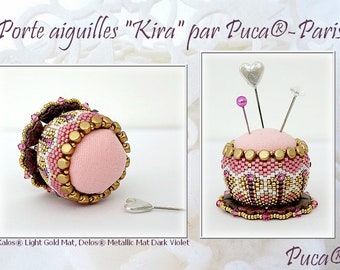 May include: A pink beaded pincushion with gold and white accents. The pincushion is shaped like a small cake and has a heart-shaped pin holder on top. The pincushion is sitting on a white surface.