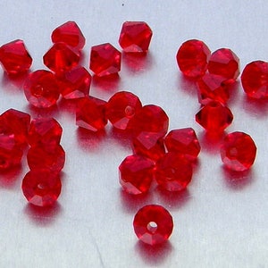 May include: A collection of faceted, red glass beads scattered on a light-colored surface. The beads have a variety of shapes, including round and multi-sided, with holes for stringing. The beads are a vibrant, transparent red.