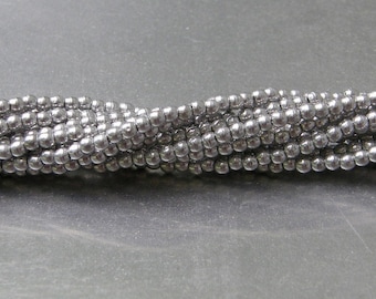 May include: Close-up of several strands of small, round, metallic gray beads. The beads are tightly strung together, creating a textured, braided appearance. The beads have a shiny, reflective surface, and the background is a solid, neutral gray.