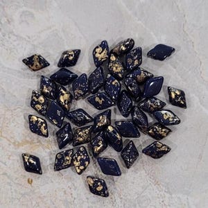 以下が含まれることがあります： 金色の斑点のある濃い青色のガラスビーズの山。ビーズはダイヤモンド型で、薄い灰色の表面に散らばっています。