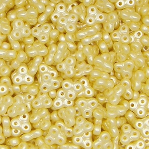 May include: A close-up view of a large quantity of small, pale yellow, plastic beads. The beads are shaped like a three-leaf clover with a hole in the center of each leaf.