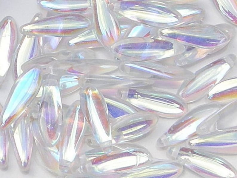 May include: A pile of iridescent white glass beads in an oval shape. The beads have a pearlescent finish and are scattered on a white surface.