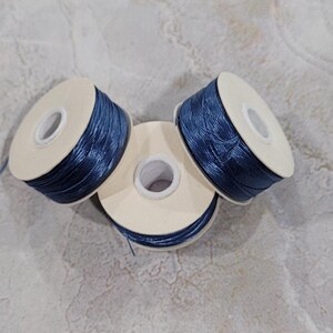 May include: Three spools of navy blue thread are arranged on a light gray surface. Each spool has a white cardboard core and is tightly wound with the thread. The thread appears to have a slight sheen.
