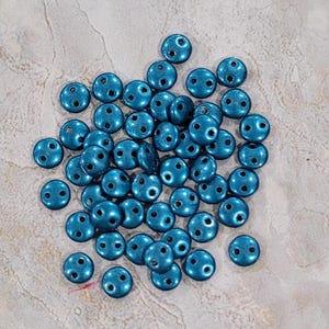 May include: A pile of teal blue metallic round beads with two holes each. The beads are arranged in a loose pile on a light gray background.