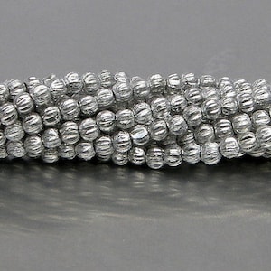 May include: A strand of silver-colored beads with a textured, ridged surface. The beads are round and have a slightly irregular shape.
