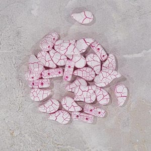 May include: A collection of white teardrop-shaped beads with pink crackle glaze. The beads are arranged in a circle on a light gray surface.