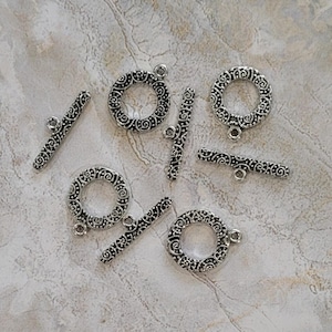 May include: Silver-toned toggle clasps with an ornate, swirling design. The clasps include circular rings and bar-shaped toggles, perfect for jewelry making. The clasps are arranged on a light-colored, textured surface.