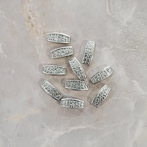 May include: A collection of silver-colored, rectangular beads with a floral design. The beads are scattered on a light gray, textured surface. The floral pattern is detailed and adds visual interest to the beads.