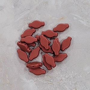 May include: A pile of small, red, ceramic beads in the shape of a teardrop. The beads have a matte finish and are arranged in a loose pile.