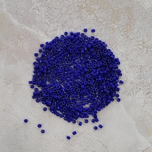 Puede incluir: Un montón de pequeñas cuentas de vidrio cilíndricas de color azul cobalto. Las cuentas están dispersas sobre una superficie texturizada de color gris claro. Estas cuentas son adecuadas para la fabricación de joyas y otros proyectos de artesanía.