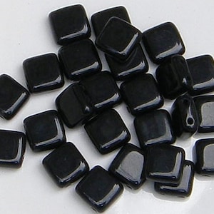 May include: A collection of glossy, square-shaped black beads. The beads are evenly sized and have rounded corners. They are scattered on a white surface, reflecting light and creating highlights.