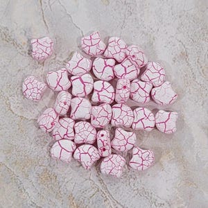 Pode incluir: Um grupo de contas de cerâmica branca com esmalte craquelado rosa. As contas têm a forma de pequenos ovais irregulares.