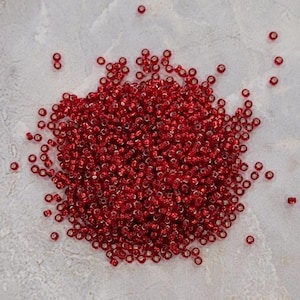 May include: A pile of small, round, red beads scattered on a light gray surface. The beads are a vibrant, glossy red color, and appear to be made of glass or a similar material. These beads are suitable for jewelry making and crafts.