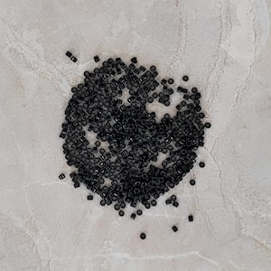 Peut inclure: Un tas de petites perles cylindriques noires dispersées sur une surface claire et grise marbrée. Les perles semblent être faites d'un matériau solide et sont de taille uniforme. L'image est un gros plan, soulignant la texture et la couleur des perles.