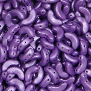 May include: A close-up view of a large quantity of small, purple, crescent-shaped glass beads. The beads have a metallic sheen and are arranged in a random pattern.