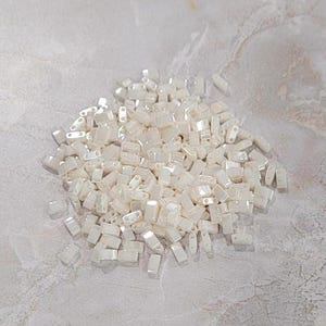 May include: A large pile of white, opaque, rectangular seed beads. The beads are arranged in a random pattern and are all the same size.