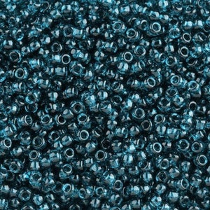 May include: A close-up view of a large quantity of teal blue glass seed beads. The beads are small and round, with a slightly faceted surface.