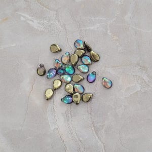 Puede incluir: Una colección de cuentas en forma de lágrima. Algunas son doradas, mientras que otras tienen un acabado iridiscente multicolor con tonos de azul, verde y morado. Las cuentas están dispersas sobre una superficie gris claro.