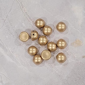 May include: A group of 10mm gold round cabochon beads with a hole in the center. The beads are scattered on a light gray surface.