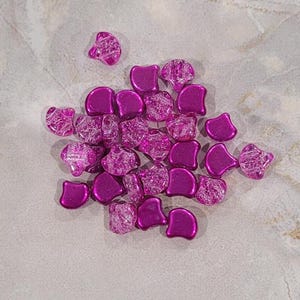 May include: A collection of shiny, fuchsia-colored beads with a unique, irregular shape. Some beads have a cracked, textured appearance, while others are smooth. The beads are scattered on a light gray surface.