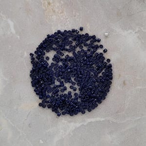 May include: A circular arrangement of small, dark blue seed beads. The beads are uniform in size and shape, with a slightly glossy finish. A few stray beads are scattered around the main cluster. The beads are likely for crafting.