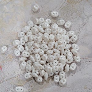 May include: A pile of white, double-holed beads with a pearlescent finish. The beads are scattered on a textured, light gray surface. The beads are small and oval-shaped, ideal for jewelry making.