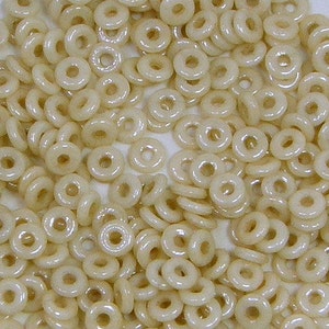 May include: A collection of small, cream-colored, donut-shaped beads. The beads are evenly distributed, filling the frame. Each bead has a smooth, glossy surface and a circular hole in the center. The beads are likely used for jewelry making or crafting.