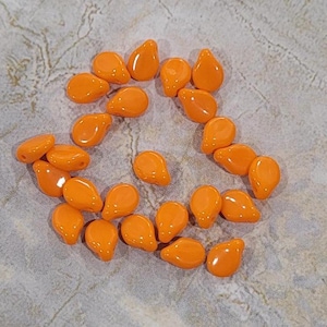 May include: A collection of orange teardrop-shaped beads. The beads are glossy and appear to be made of glass or a similar material. They are scattered on a textured, light gray surface.