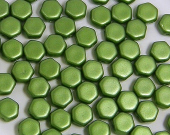 May include: A close-up of a group of green hexagonal glass beads. The beads have a matte finish and are scattered on a white surface.