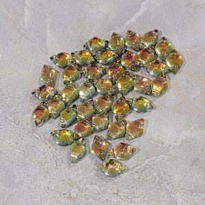 May include: A collection of iridescent, teardrop-shaped beads in shades of gold, green, and bronze. The beads are scattered on a light gray, textured surface. These beads are suitable for jewelry making and crafting projects.