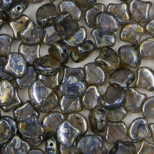 May include: A close-up of a group of small, iridescent glass beads in a dark blue and gold color. The beads are shaped like a teardrop with a flat back and a rounded front. The beads are scattered on a white surface.