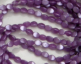 Könnte beinhalten: Nahaufnahme eines Strangs aus lila, oval geformten Perlen. Die Perlen haben eine perlmuttartige Oberfläche mit einem helleren, fast weißen Glanz auf einer Seite. Die Perlen sind aufgefädelt und erzeugen ein fließendes, organisches Muster. Ideal für die Schmuckherstellung.