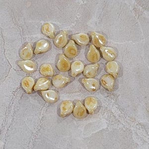 May include: A collection of teardrop-shaped beads with a creamy white base and golden-brown marbling. These small beads are scattered on a textured, light gray surface, ideal for jewelry making or craft projects.