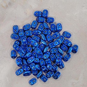 Può includere: Un mucchio di perline ovali blu metallizzato, ciascuna con due fori. Le perline sono sparse su una superficie grigio chiaro, mostrando la loro finitura lucida. Adatte per la creazione di gioielli.