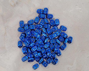 Puede incluir: Un montón de cuentas ovales de color azul metálico, cada una con dos agujeros. Las cuentas están dispersas sobre una superficie gris claro, mostrando su acabado brillante. Adecuadas para la creación de joyas.