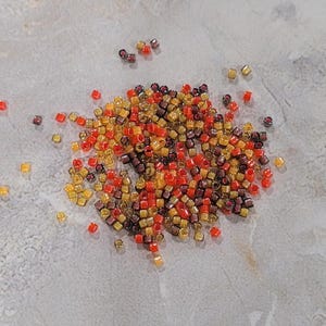 May include: A pile of small, square, translucent beads in shades of red, orange, yellow, and brown. The beads are scattered on a light gray surface.
