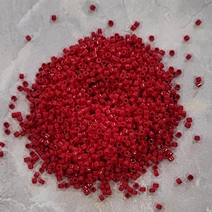 May include: A collection of small, red, cylindrical beads scattered on a light grey surface. The beads are uniform in size and shape, with a glossy finish. These beads are likely used for jewellery making or other craft projects.