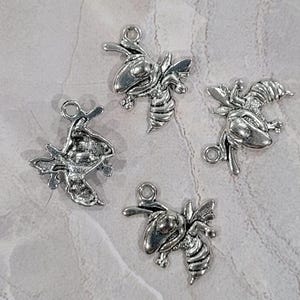 May include: Four silver-colored bee charms with detailed wings and striped bodies. Each charm has a small loop at the top for attaching to jewelry or other crafts. The charms are scattered on a light gray, textured surface.