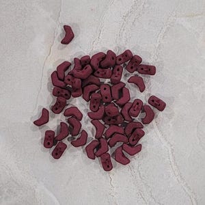 May include: A pile of small, burgundy-colored ceramic beads in the shape of a crescent moon. The beads are scattered on a white surface.