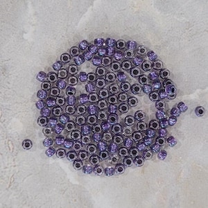May include: A close-up of a pile of small, iridescent purple glass beads. The beads are round and have a metallic sheen.