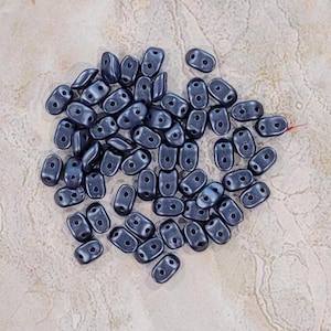May include: A collection of dark grey seed beads, also known as rocailles, in a teardrop shape. The beads are arranged in a loose pile on a light beige surface.
