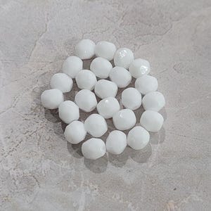 Op de afbeelding: Een groep witte glazen kralen met een gefacetteerde, diamantvormige vorm. De kralen zijn in een losse cirkel op een lichtgrijs oppervlak gerangschikt.